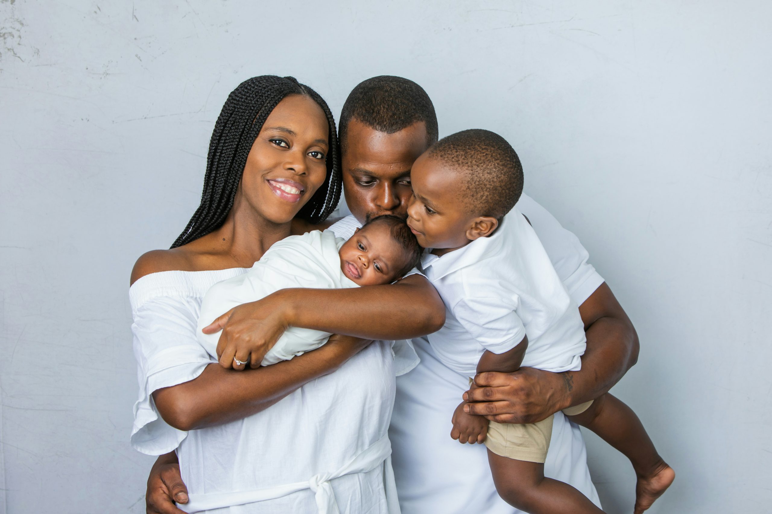 A young African-American family in the studio with two sons, one who is a toddler and one who is a newborn boy and dad and brother are kissing the new baby boy