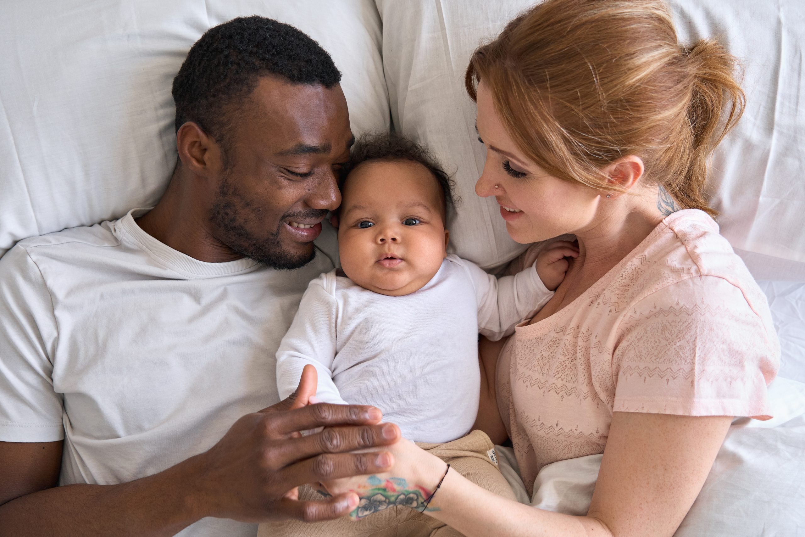 Mixed race young family couple lying in bed bonding with cute infant girl. Happy multiracial parents relaxing waking up in the morning playing with small baby daughter in bedroom at home, top view.
