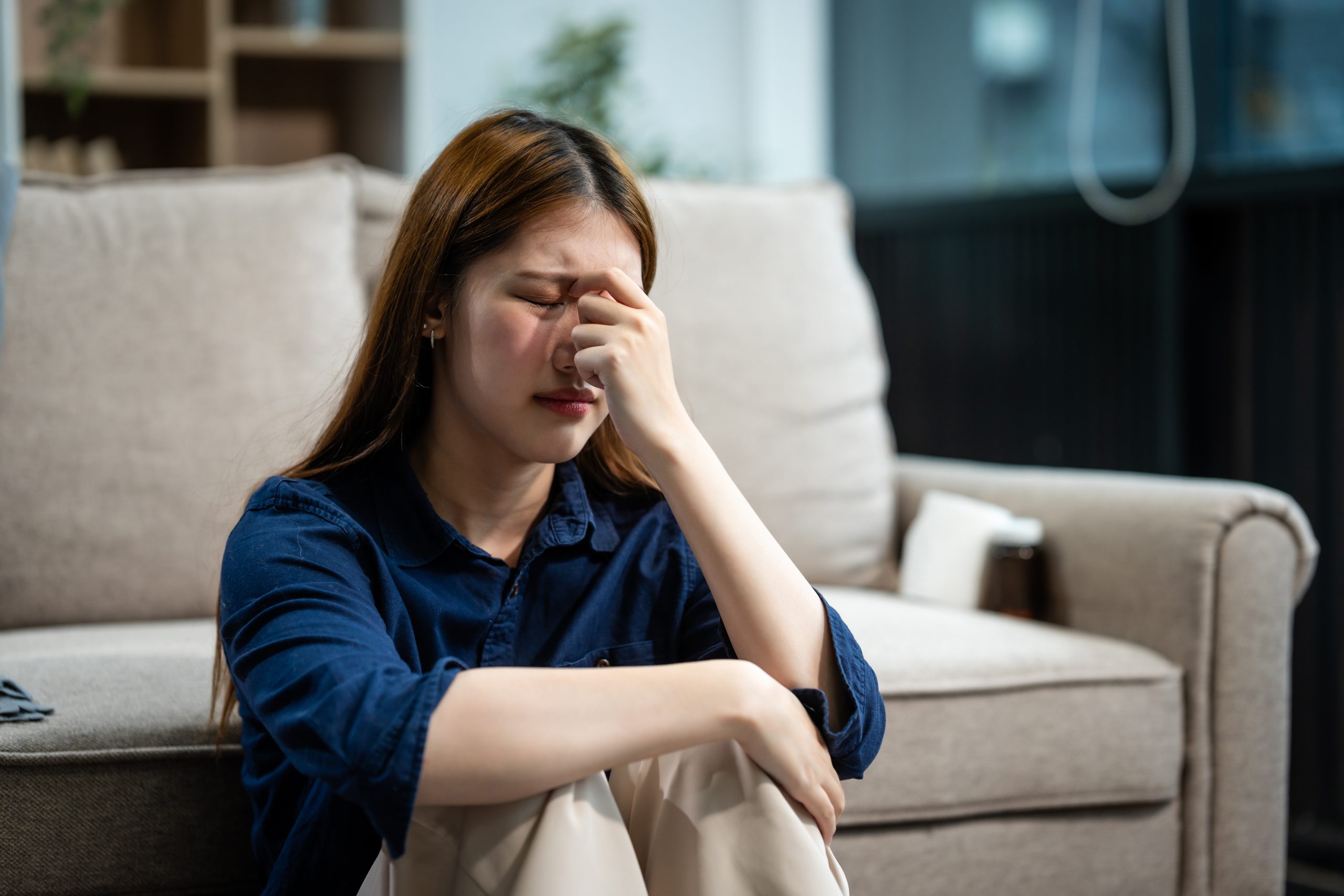 A young Asian woman sits on a sofa in her living room, feeling sick with mental health diseases. She struggles with depression, anxiety, and other conditions, seeking support and solace at home.
